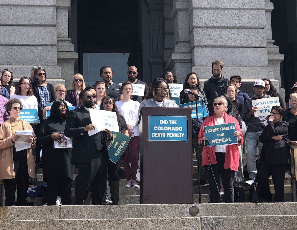 A group of people stands outside of a government building with signs that read "End the Colorado Death Penalty"