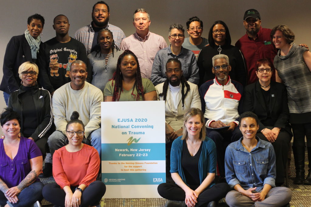 A group of about 20 people gather around a sign that reads "EJUSA 2020 Convening on Trauma"