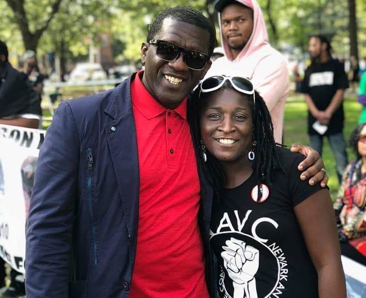 Lakeesha Eure, a black woman with locs, stands in a park smiling with a community member. The man is wearing sunglasses, a red shirt and blazer, and is taller than Lakeesha.
